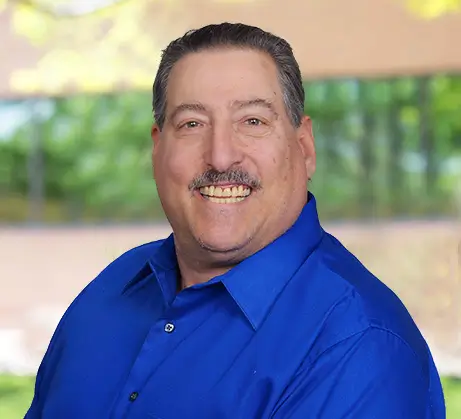 Headshot of Michael, a middle aged man smiling with short dark hair and a mustache, wearing a blue button-down shirt.